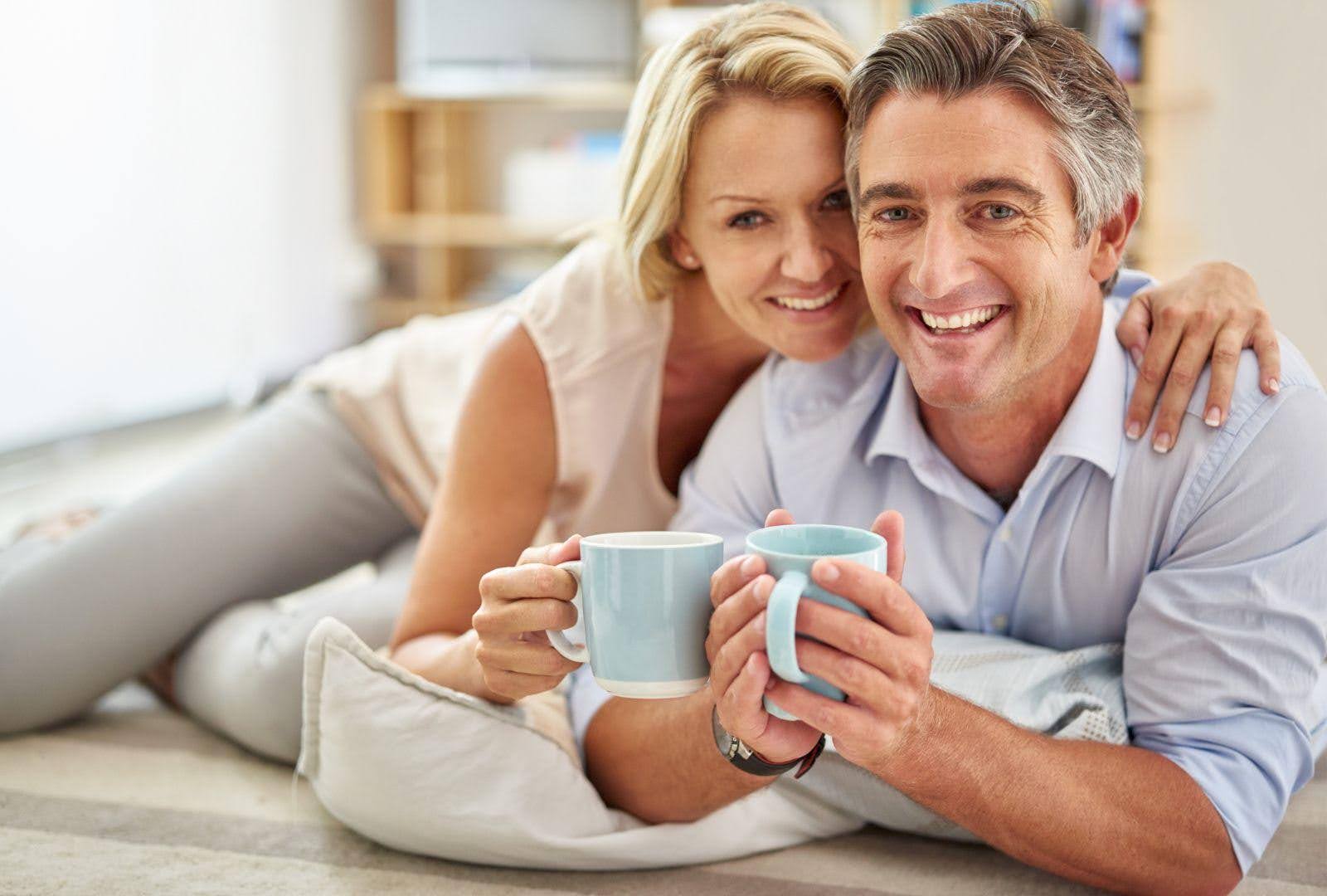 A middle-aged couple relaxing at home with cups of coffee, smiling warmly at the camera. They exude a sense of closeness and contentment, illustrating a positive moment in their adjustment to an empty nest.